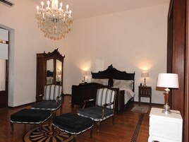 Een hotelkamer met een groot bed, twee fauteuils, een houten dressoir, een spiegel en een kroonluchter.