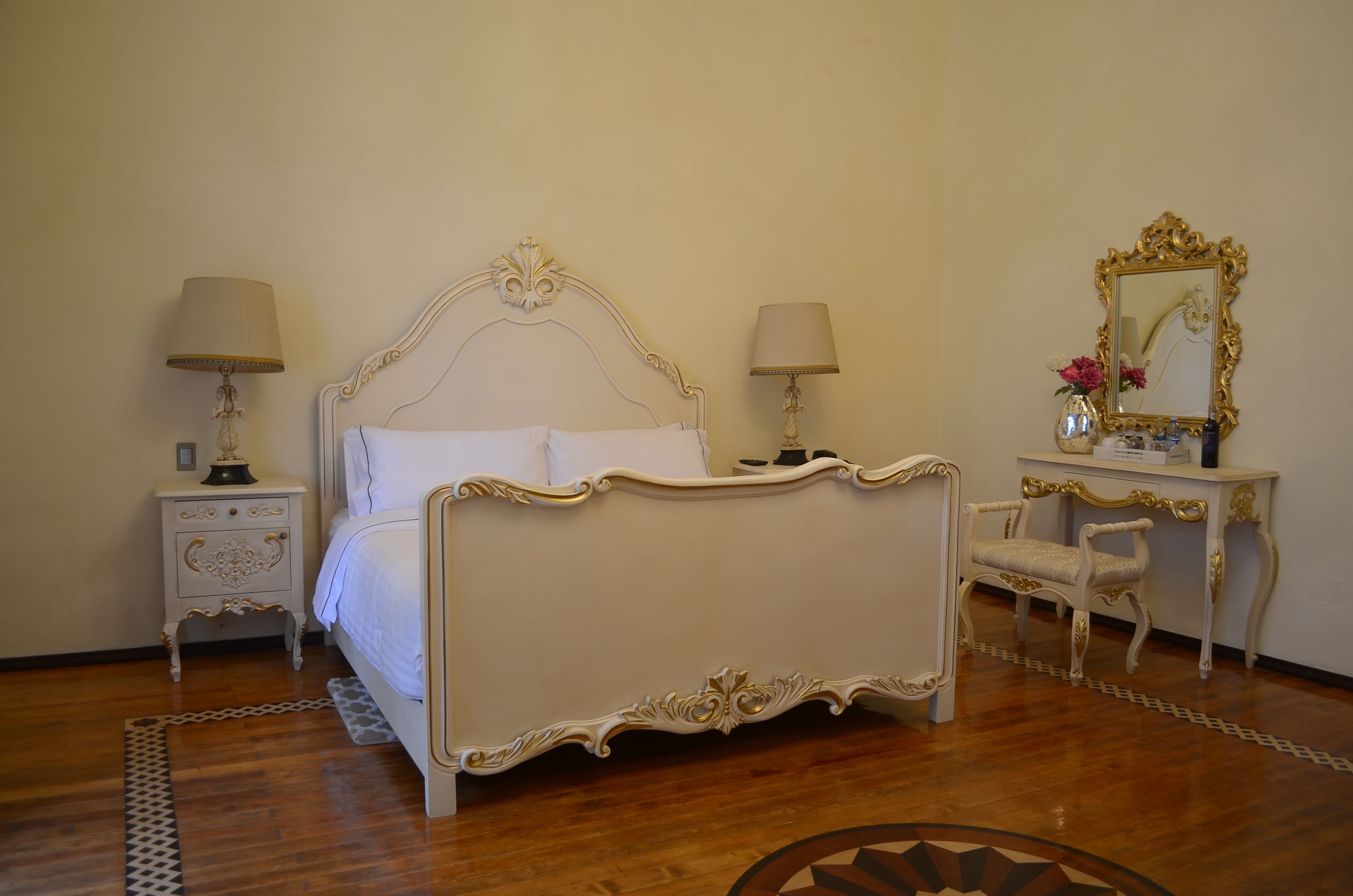 A classic four-poster bed with ornate detailing, flanked by two bedside tables with lamps, a mirror, and a chair.