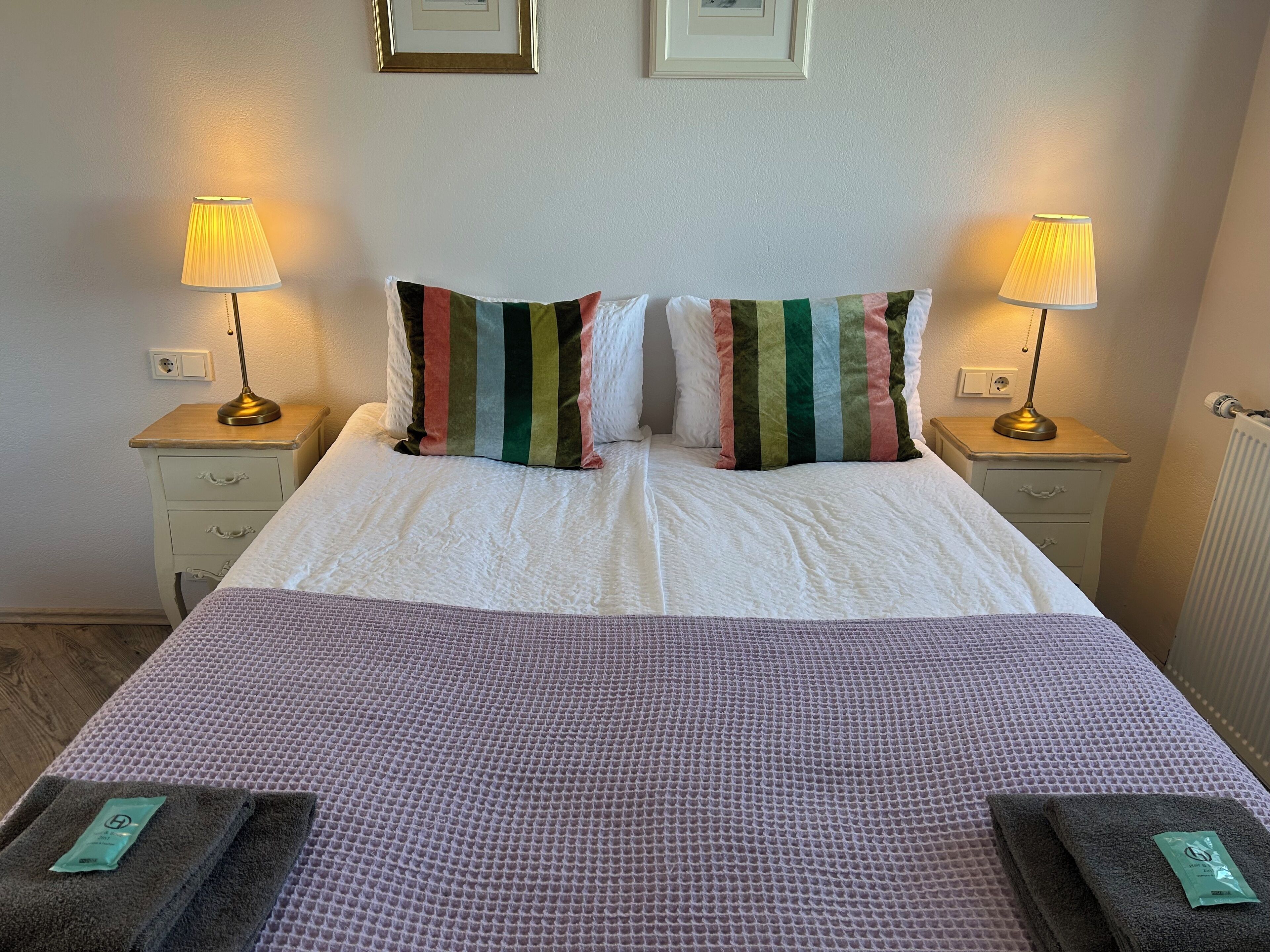 A neatly made bed with a white duvet and striped pillows, two bedside lamps, a wooden nightstand with drawers, and a textured purple blanket.