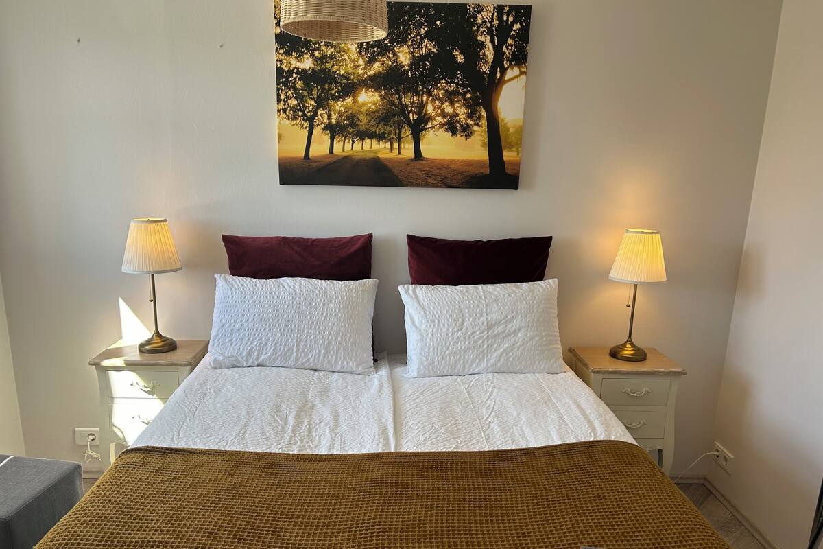A neatly made bed with white linens, two pillows, and a brown blanket. There are two bedside lamps and a painting of trees on the wall.