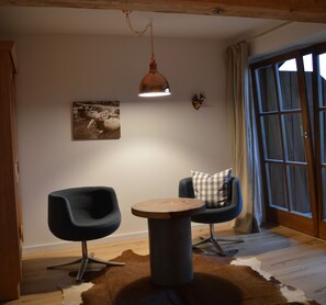 Una habitación con dos sillas modernas, una mesa redonda de madera, una cabeza de ciervo montada, un cuadro enmarcado y un amplio ventanal con cortinas.