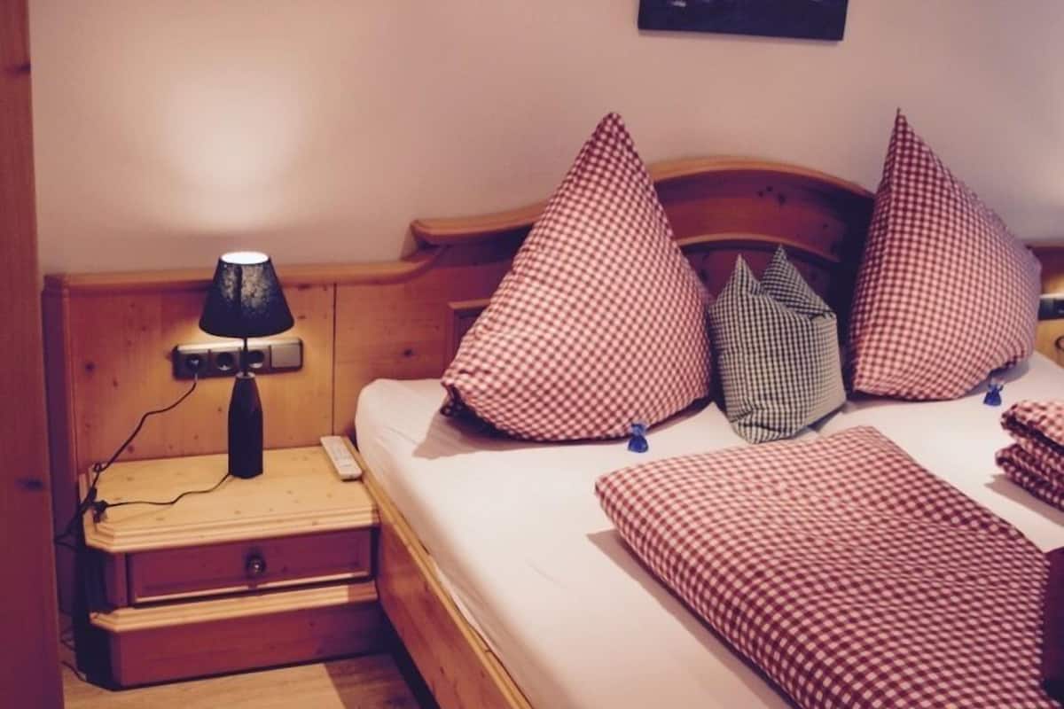 A bedroom with a wooden bed, checkered pillows, a bedside lamp, and a mountain landscape picture on the wall.