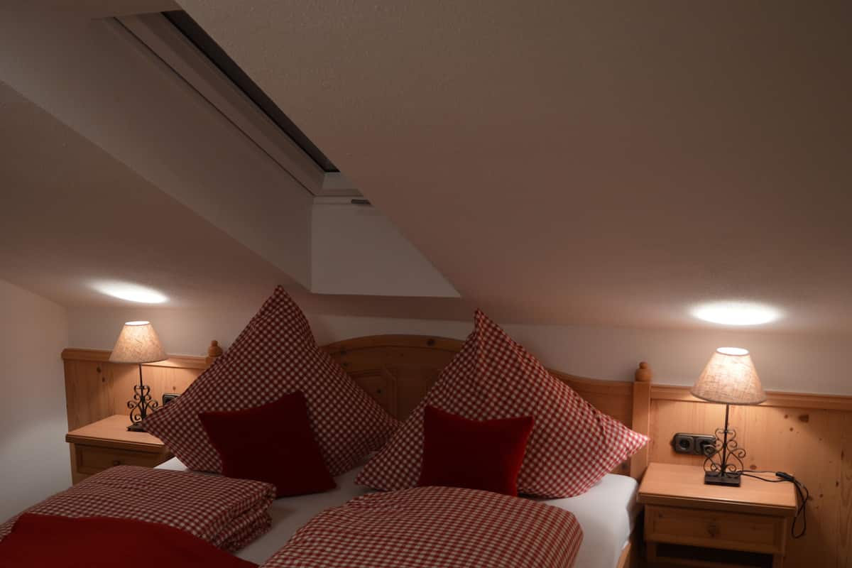 A bedroom with a sloped ceiling, two bedside tables with lamps, a bed with checkered bedding and red pillows, and a wooden headboard.