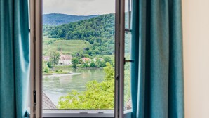 Una ventana con vista a un río y follaje, cortinas entreabiertas, y una cama con almohadones.