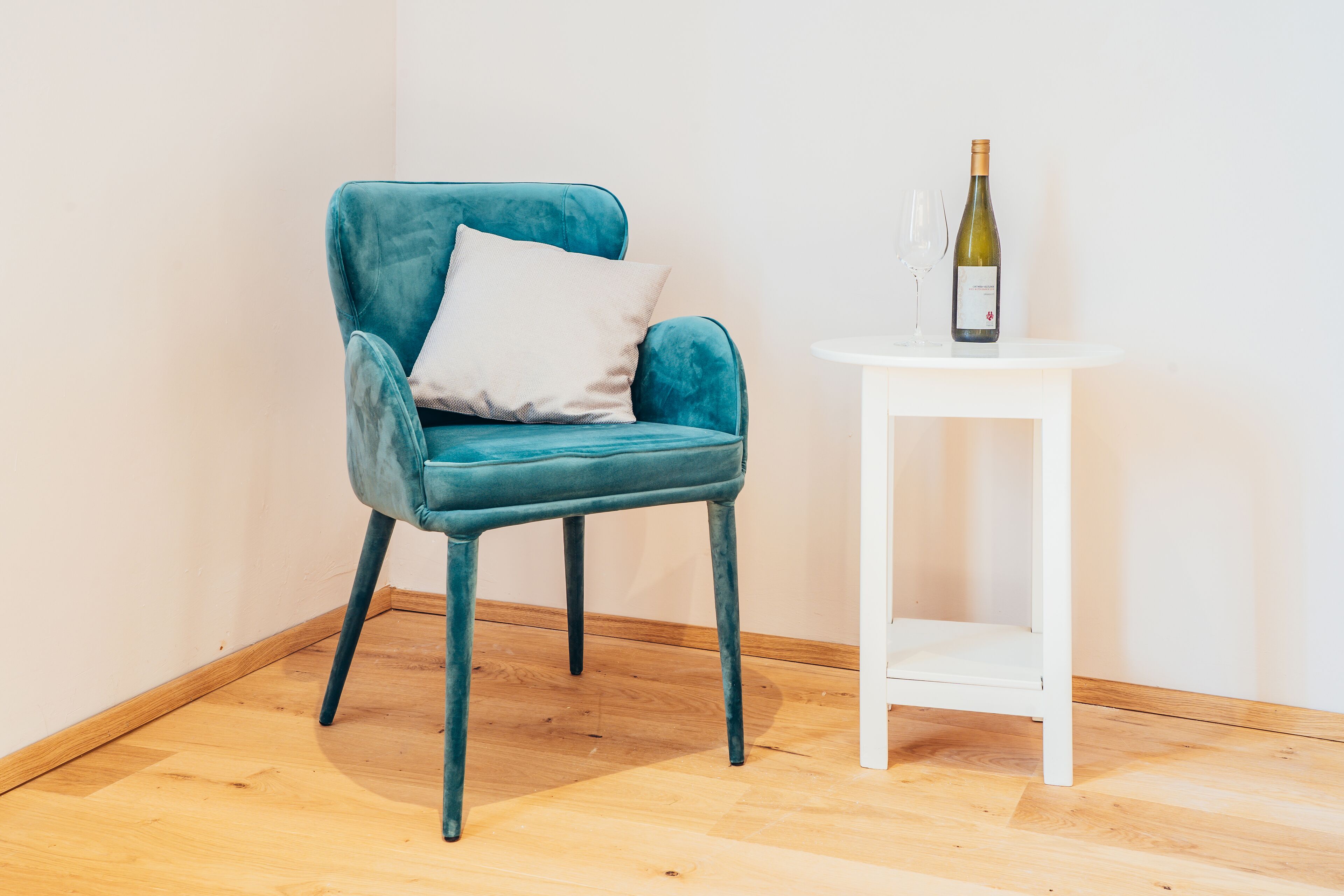 A teal armchair with a white pillow, a white side table, and a bottle of wine on the table.