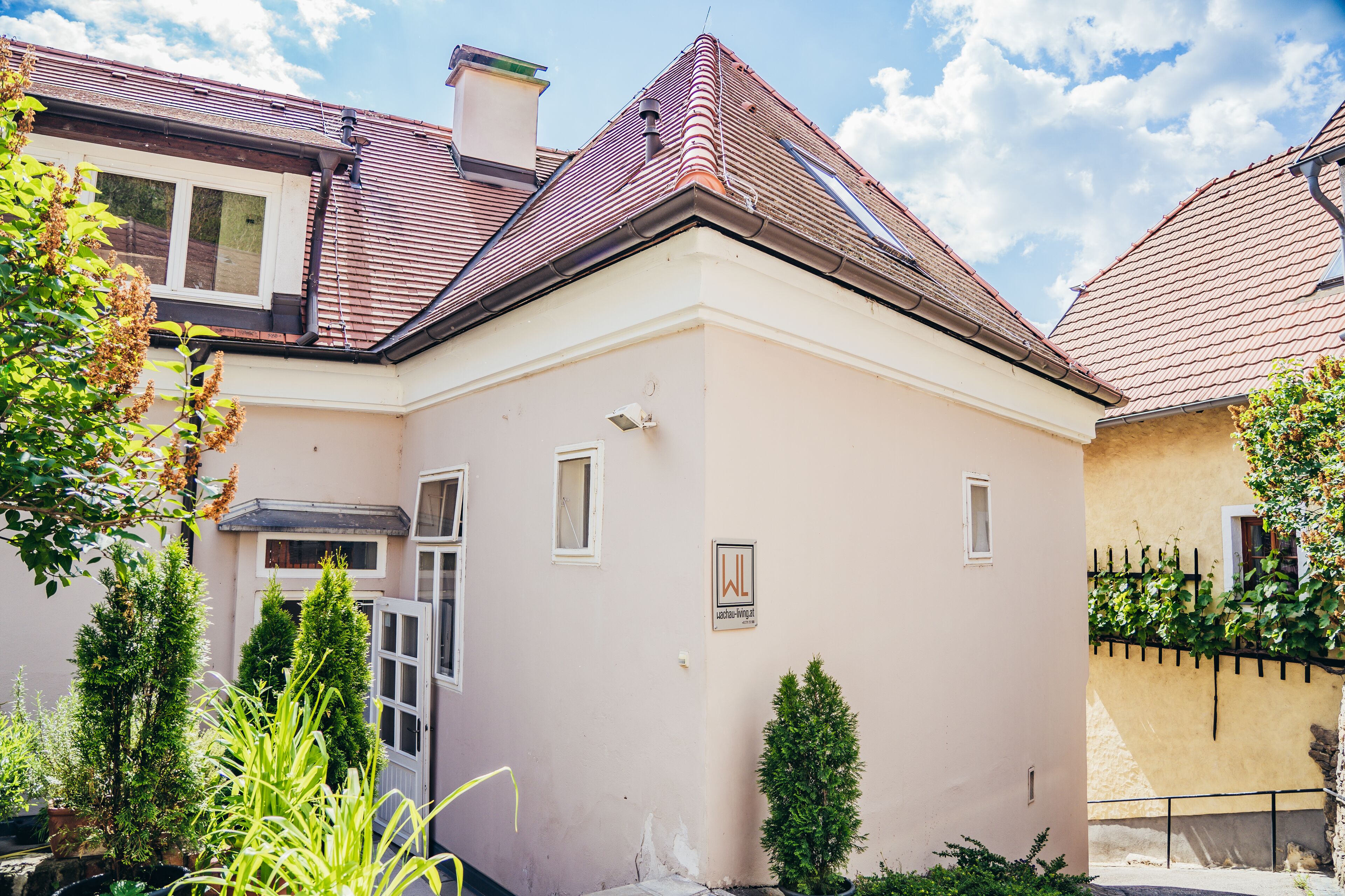 A two-story house with a red-tiled roof, a beige facade, and a small garden area with potted plants.
