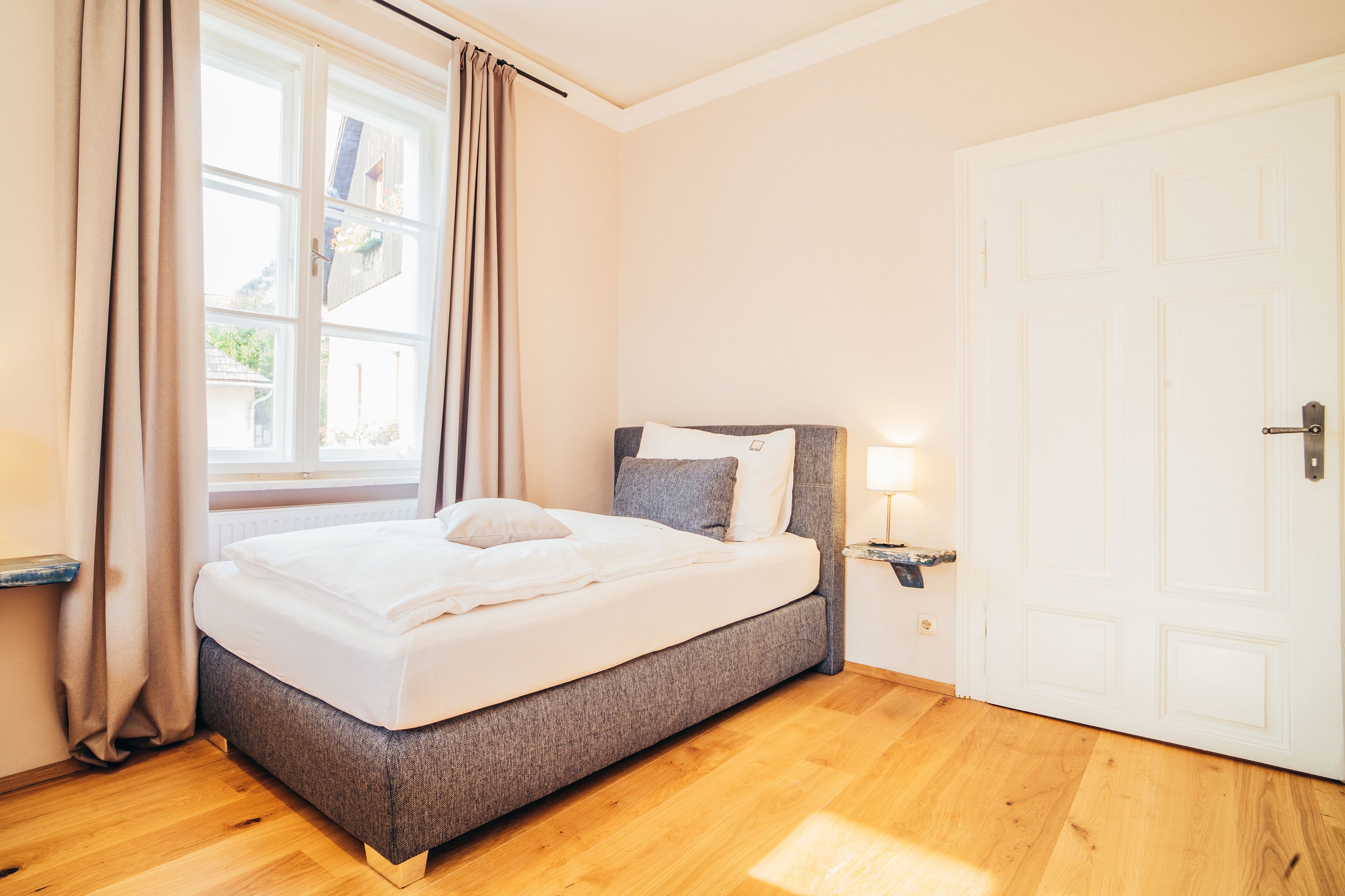 Un dormitorio con cama, ventana con cortinas, puerta y mesita de noche con lámpara.