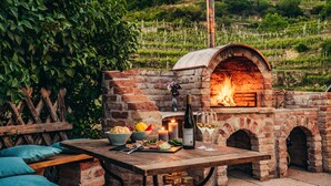 Un horno de ladrillo al aire libre con fuego, una mesa con comida y bebidas, y un banco.