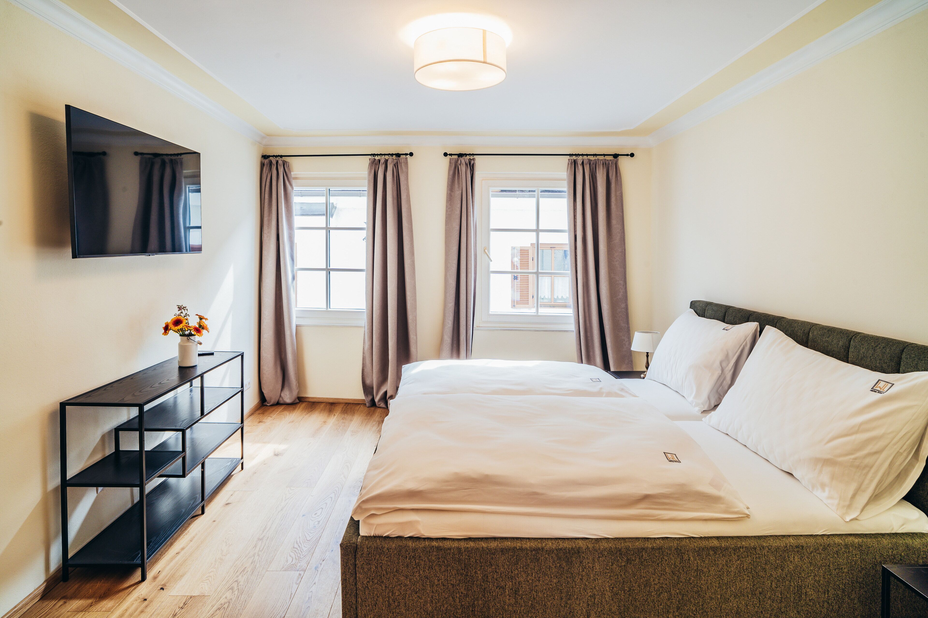 A hotel room with a bed, a TV mounted on the wall, a console table with a vase of flowers, and a window with curtains.