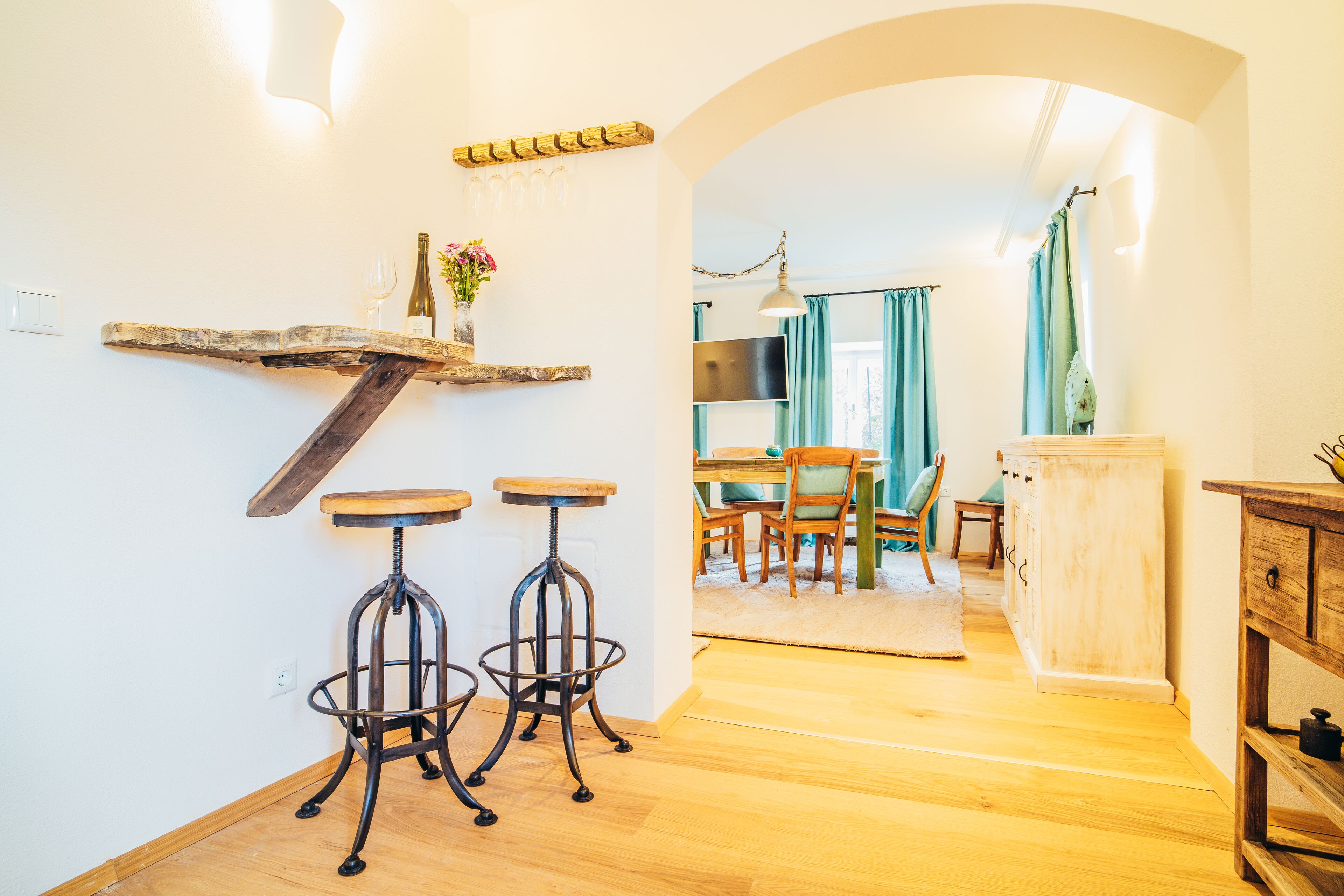 A kitchen with wooden bar stools, a hanging light fixture, and a wooden shelf with a wine bottle and glasses.