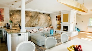 Una cocina moderna con lavavajillas integrado, una zona de comedor con mesa y sillas blancas, un sofá con cojines y una pared decorativa de piedra.
