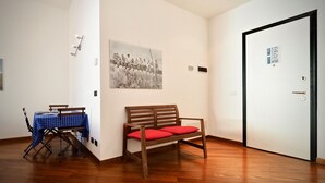 Una stanza con pavimento in legno, un tavolo a scacchi blu, una panca di legno con cuscini rossi, una porta con cornice nera e una fotografia in bianco e nero appesa al muro.