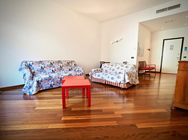 Een kamer met een houten vloer, een gestreept sofa, een bed met een bedspreide met patroon, een klein rode tafeltje en een houten kast.