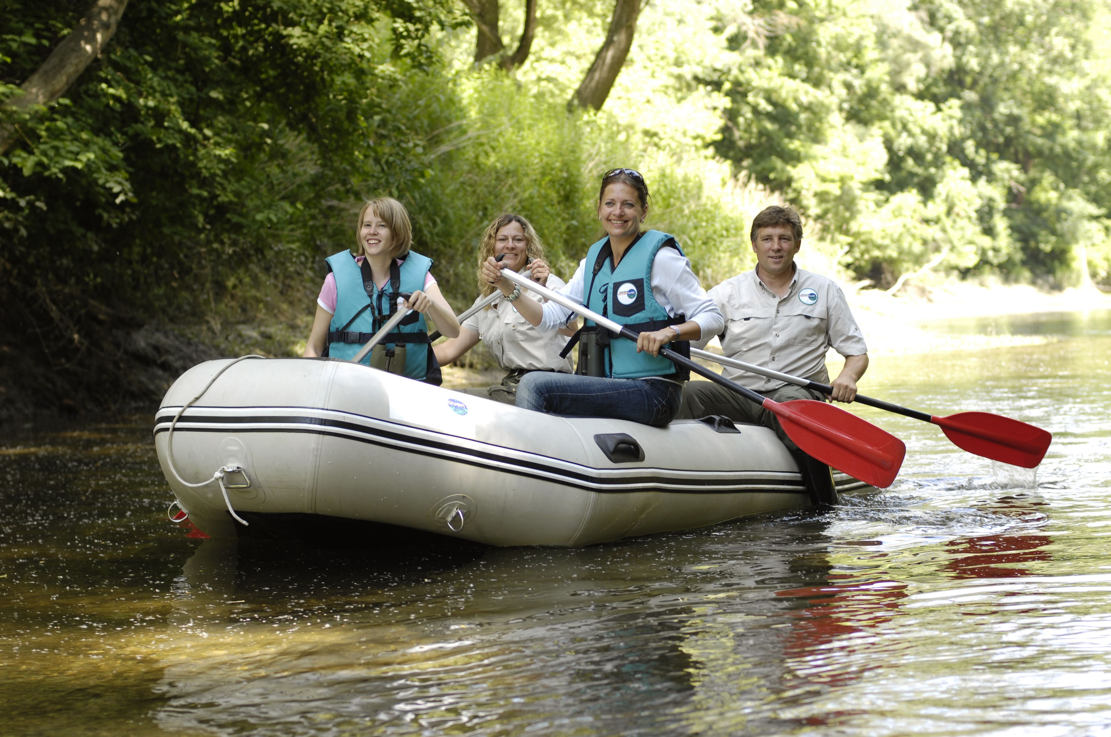 A group of people in a white inflatable raft on a calm river surrounded by dense greenery.