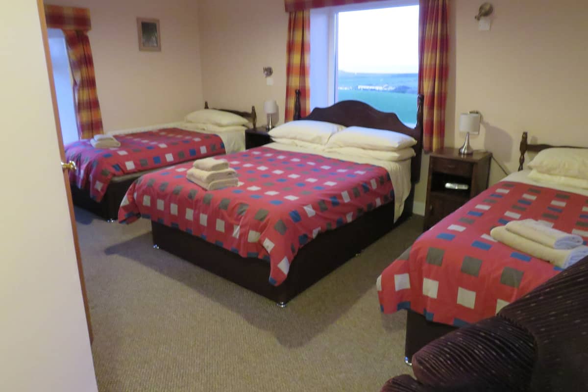 Habitación de hotel con tres camas, cada una con colcha a cuadros rojos y blancos y toallas dobladas con cuidado. La habitación tiene un ventanal con cortinas, una mesita de noche con lámpara y vistas al mar.