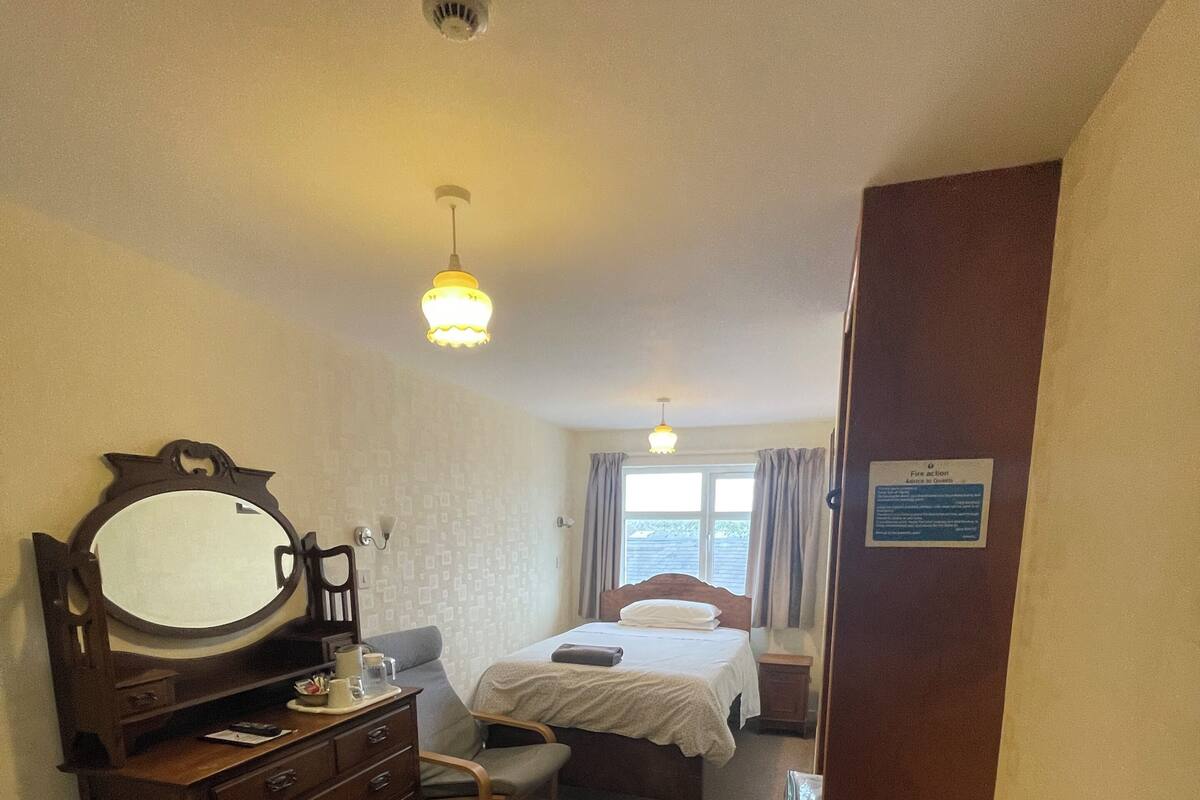 Habitación de hotel con cama, cómoda, espejo, escritorio, silla, ventana con cortinas y puerta con cartel.