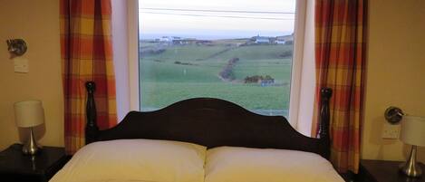 A bed with white pillows and a red and black patterned bedspread, flanked by two bedside tables with lamps, in a room with plaid curtains and a view of a green landscape through a window.