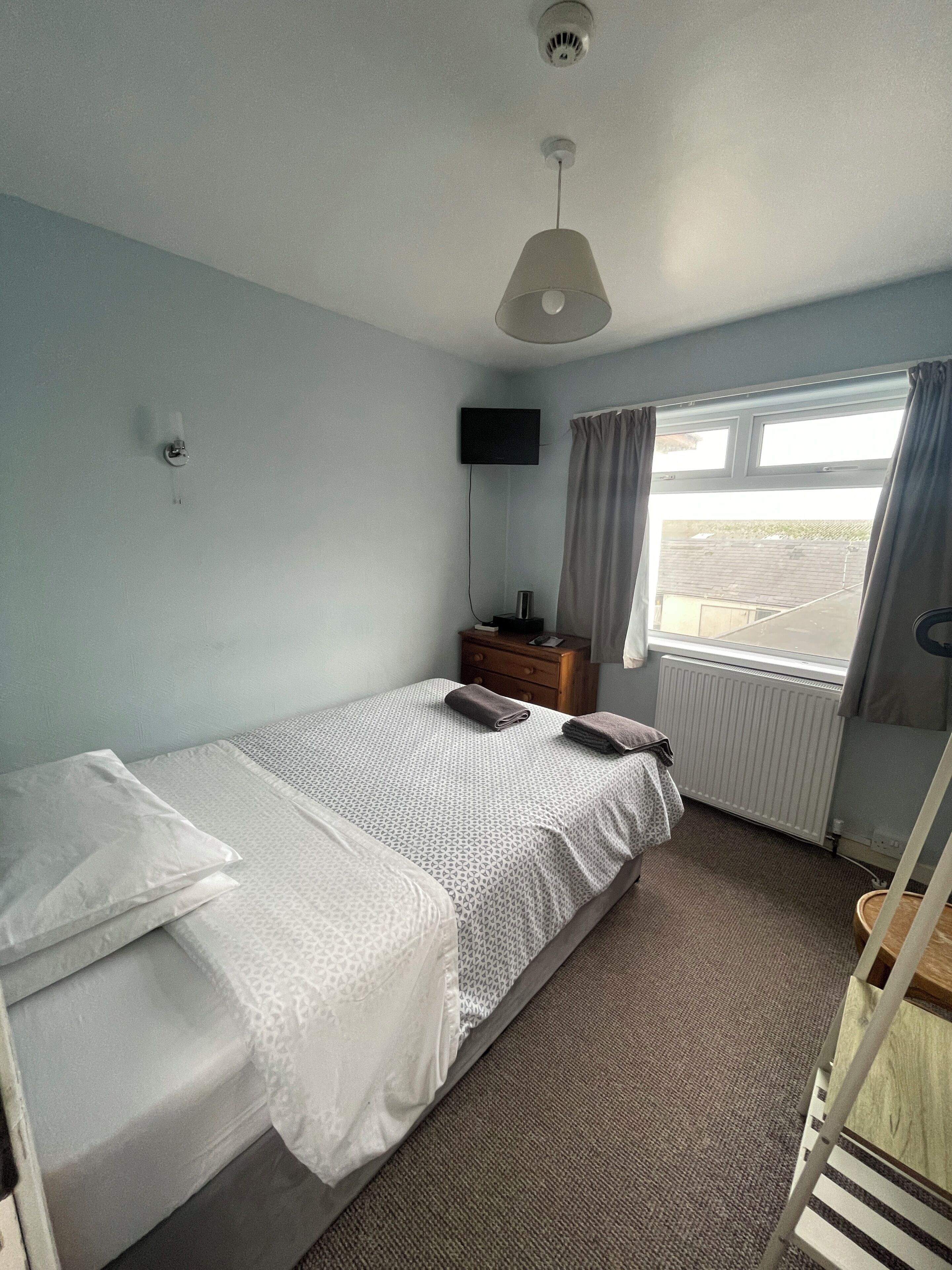 A single bed with a patterned bedspread, a bedside table with a lamp, a TV mounted on the wall, and a window with curtains.