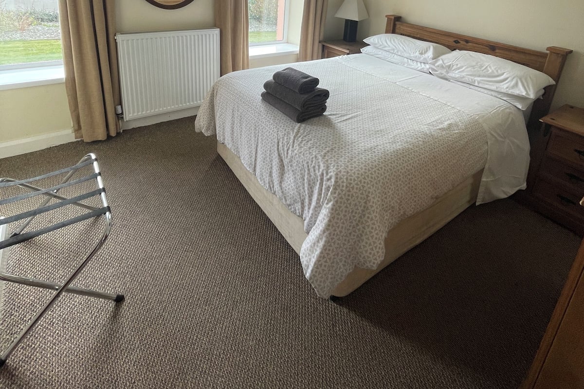 Habitación de hotel con cama, una silla, mesita de noche, lámpara, ventana con cortinas, radiador y vistas al exterior.