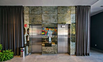 Elevador com painel de controle e instruções de emergência, emoldurado por cortinas e uma parede de pedra.
