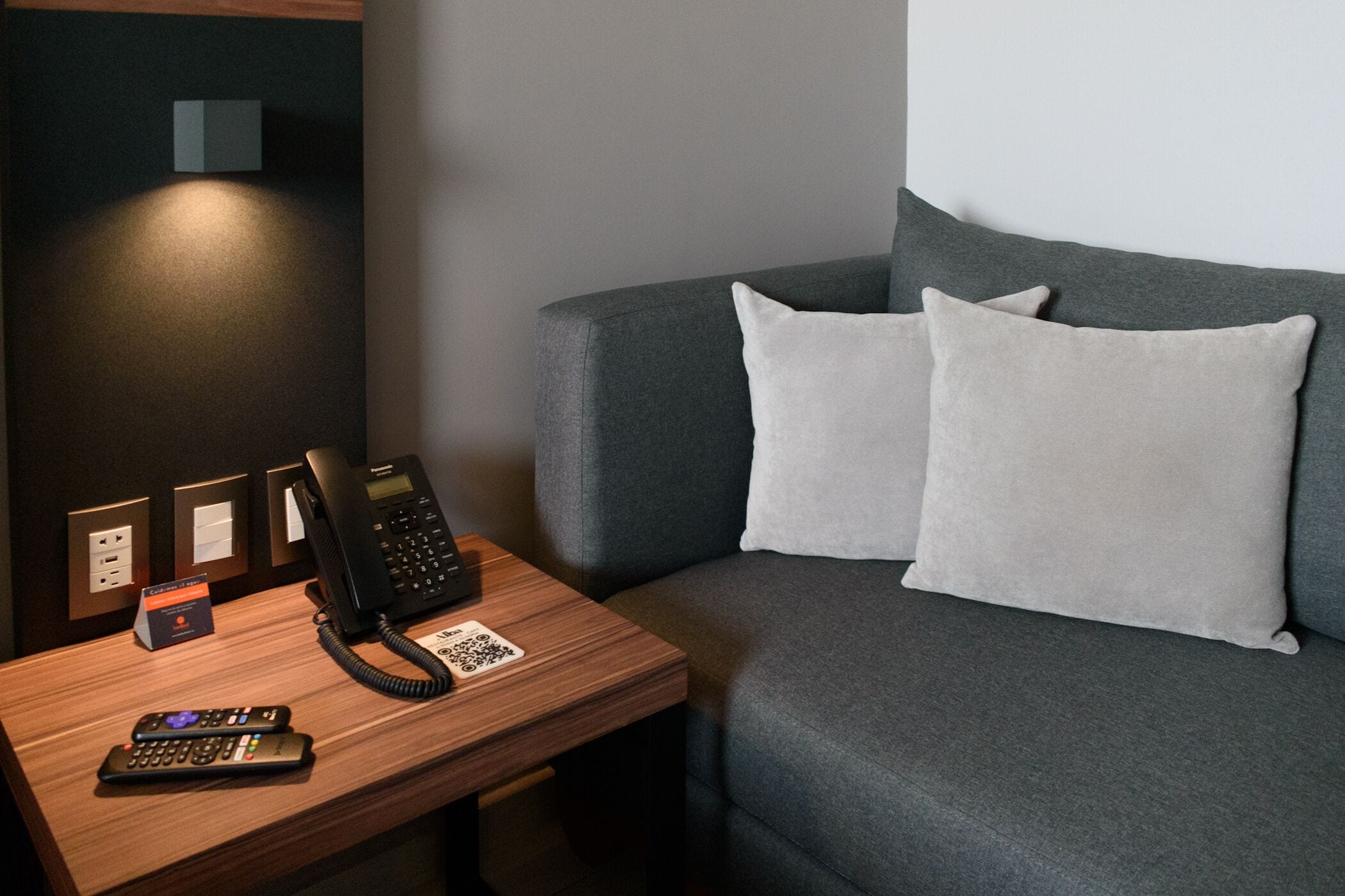 A hotel room with a wooden desk, a black telephone, a card reader, and a grey sofa with two pillows.