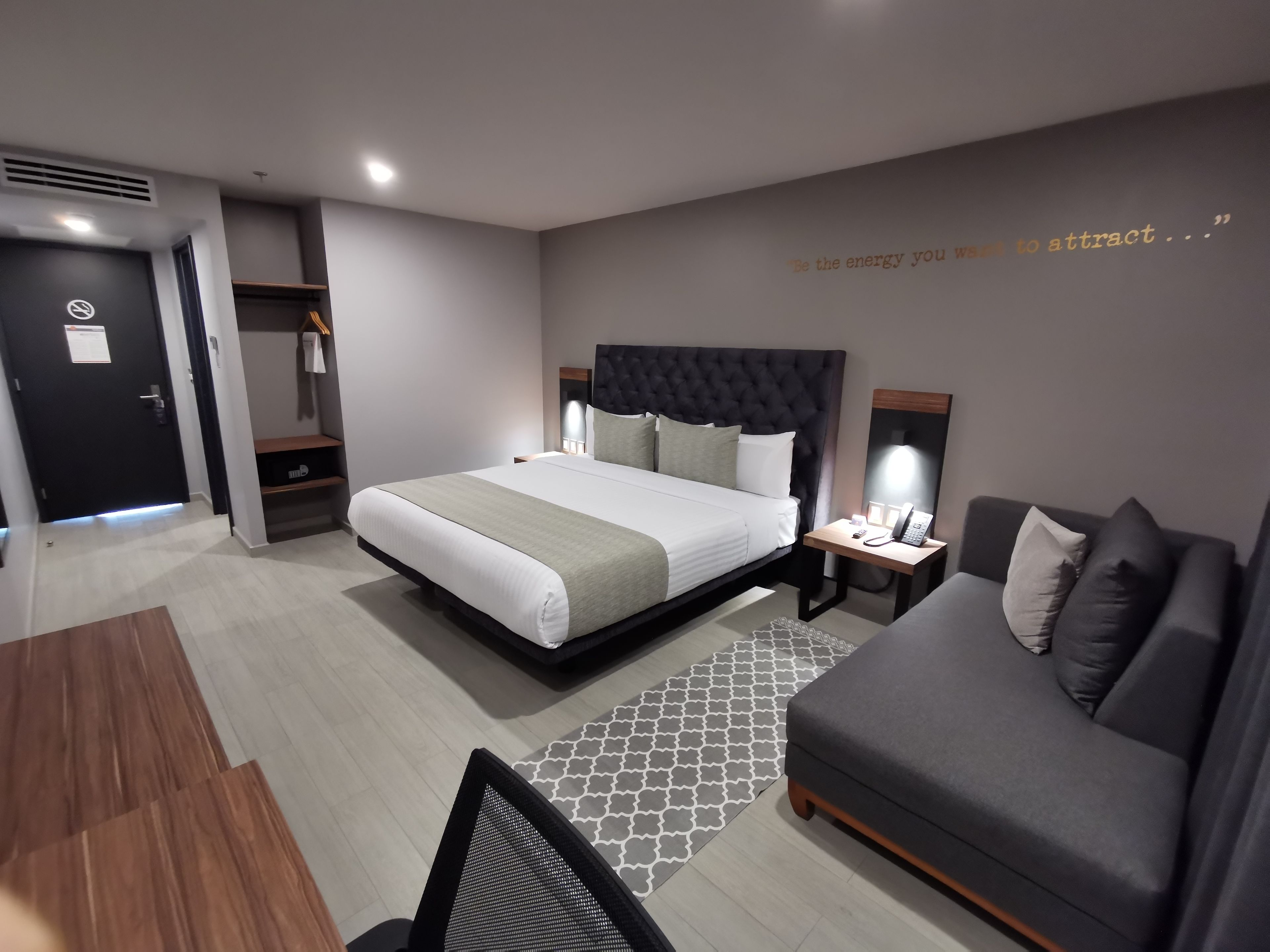 A modern hotel room with a large bed, a bedside table with a lamp, a sofa, and a patterned rug.