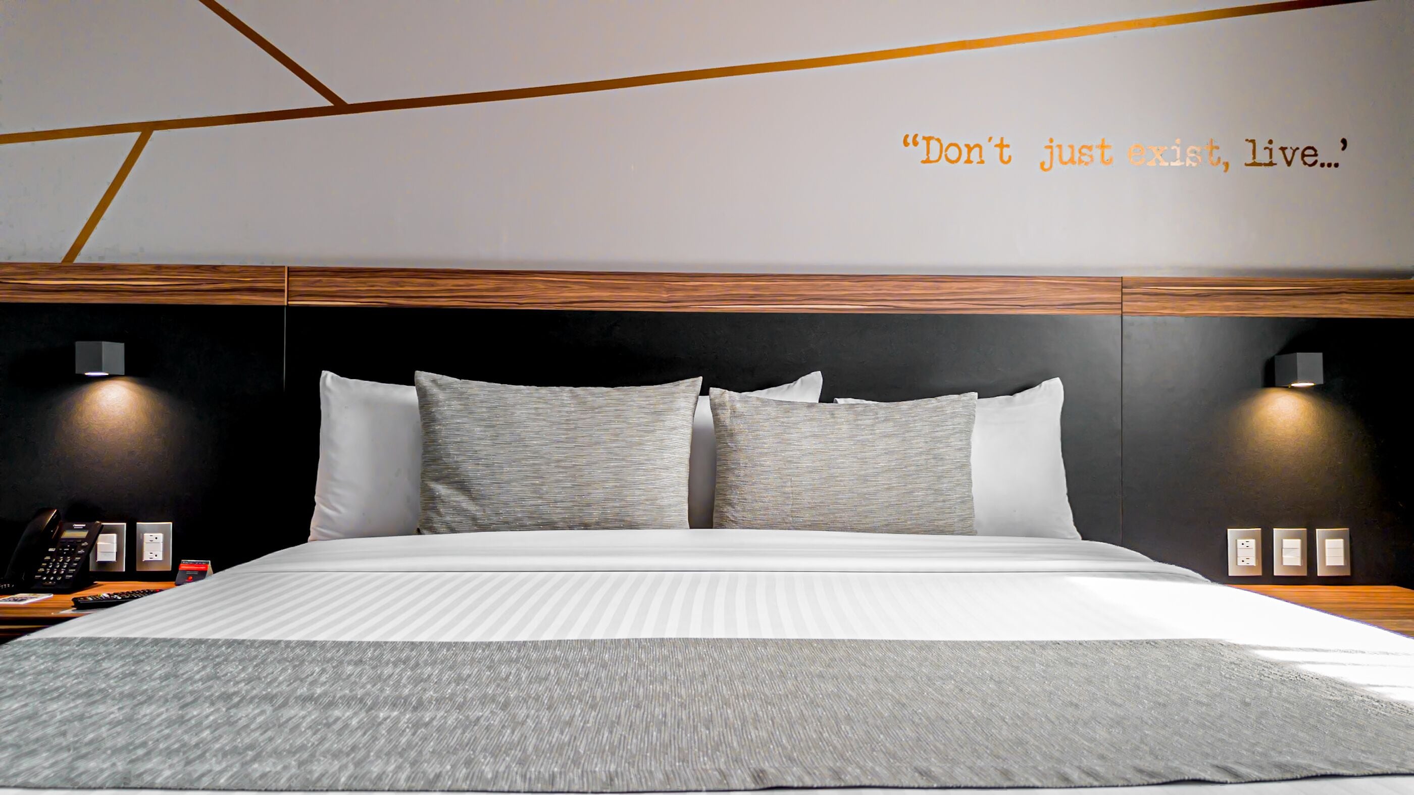 Une chambre d’hôtel moderne avec un grand lit, une tête de lit en bois, deux lampes de chevet, un téléphone et une citation affichée sur le mur : « Ne vous contentez pas d’exister, vivez… ».