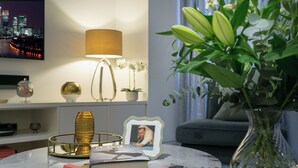 Una sala de estar con una mesa de centro redonda, un jarrón con flores, una foto enmarcada, una lámpara y un televisor que muestra un paisaje urbano.