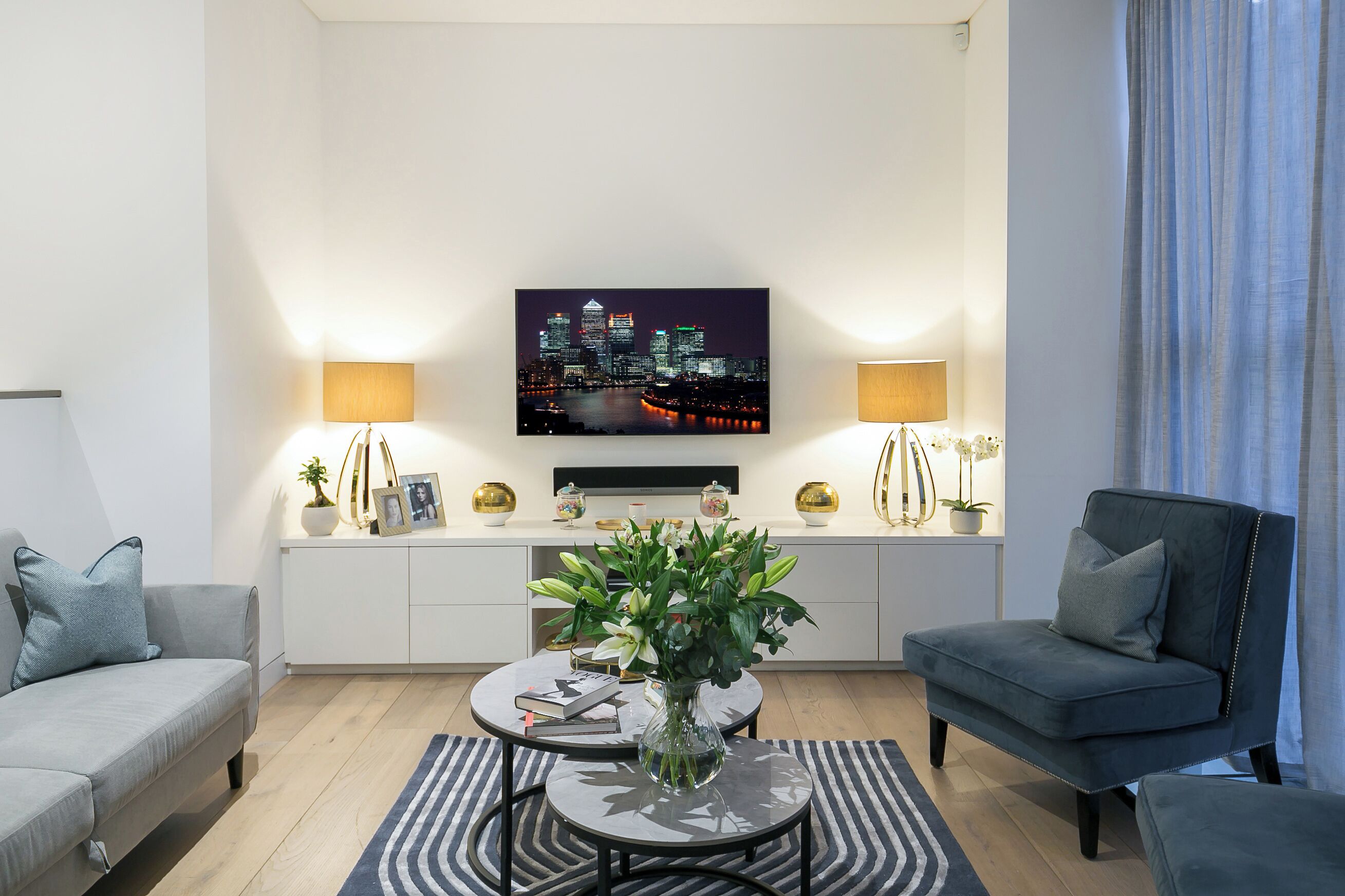 Una sala moderna con televisor de pantalla plana, dos lámparas, un sofá, una silla, una mesa de centro con un jarrón de flores y una estantería.