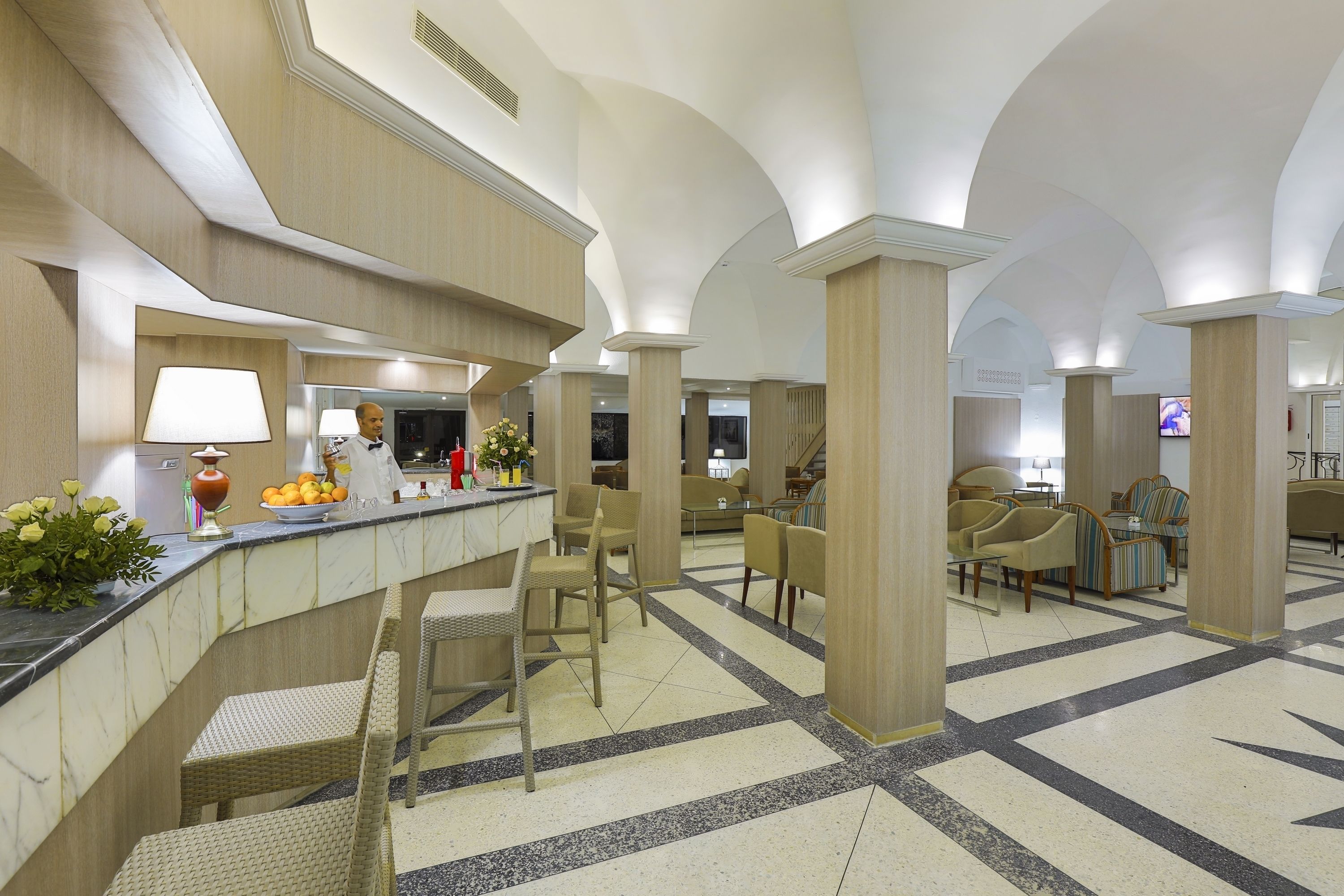 A hotel lobby with a marble reception desk, wicker chairs, and a tiled floor.
