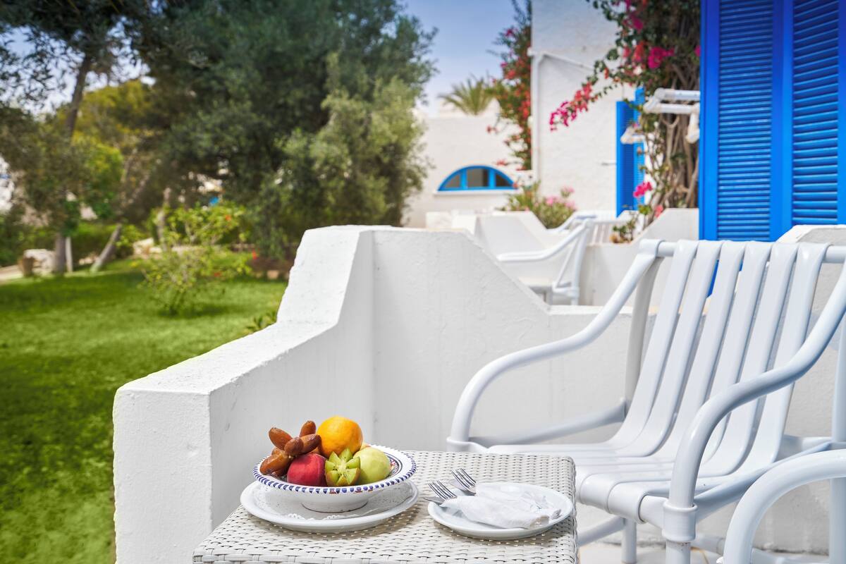 A white outdoor seating area with a table set for a meal, including a bowl of fruit and a plate of food.