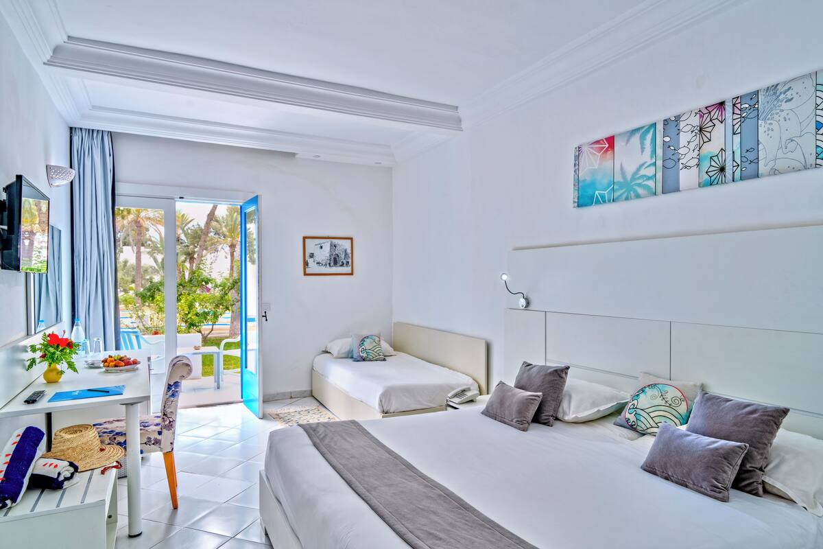 A hotel room with a bed, a desk with a computer, a chair, a window with curtains, and a painting on the wall.