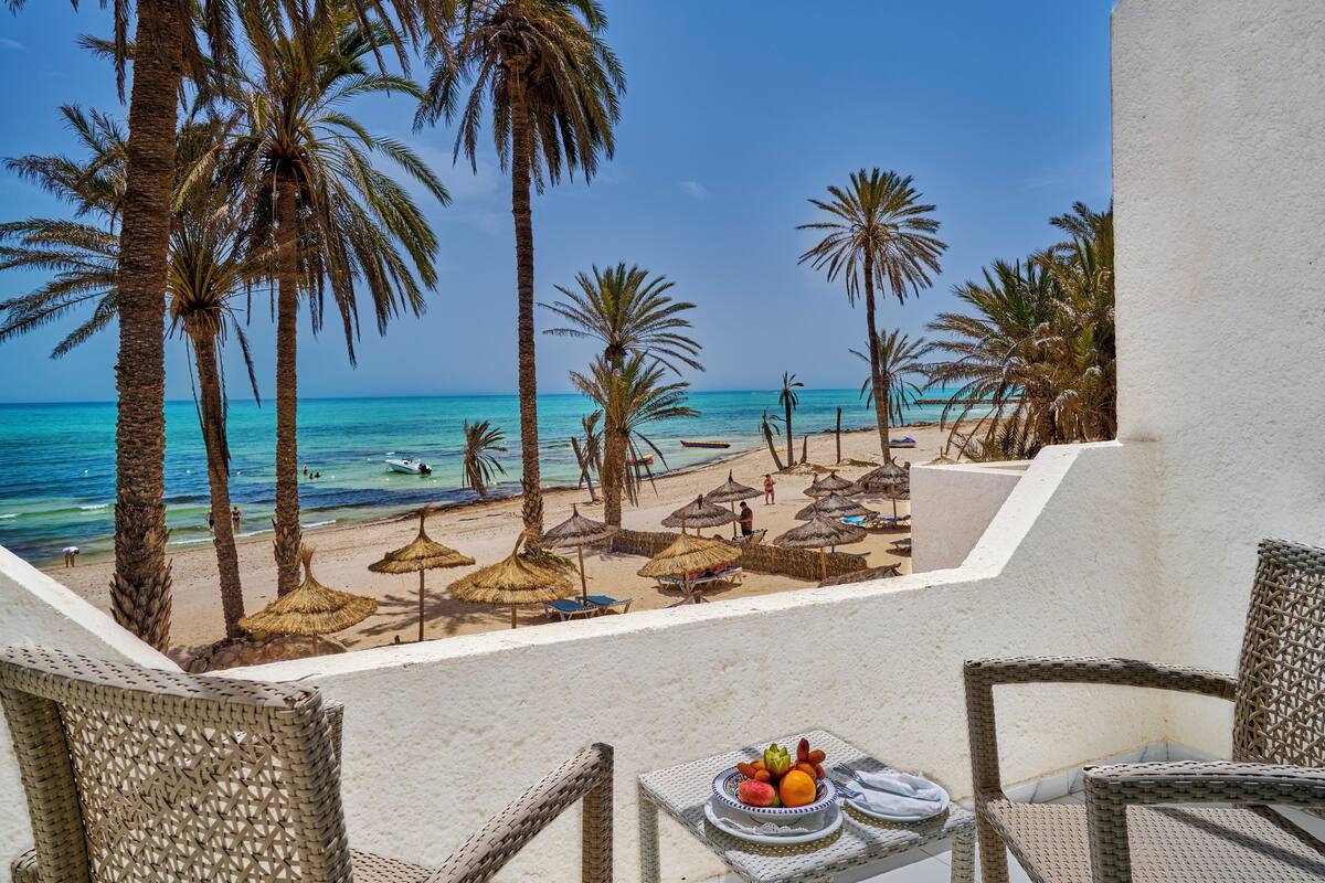 A beach view with palm trees, a table with fruit, and a chair.