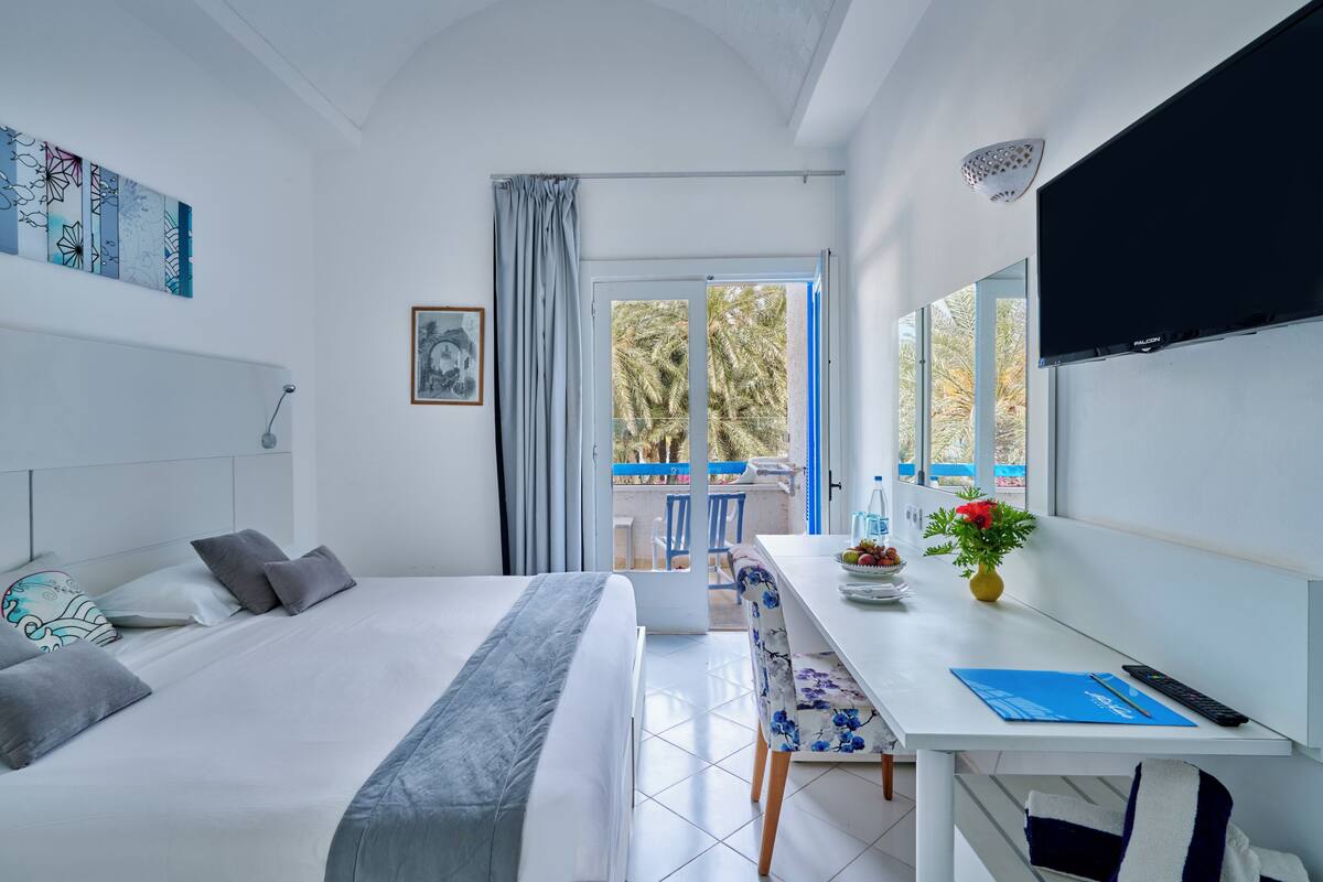A modern hotel room with a bed, a desk, a TV, and a balcony with a view of palm trees.