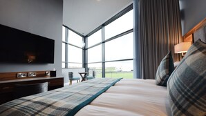 A modern hotel room with a large bed, a flat-screen TV, a desk with a lamp, and a view of a golf course through large windows. - Golflinks Hotel (Portrush)