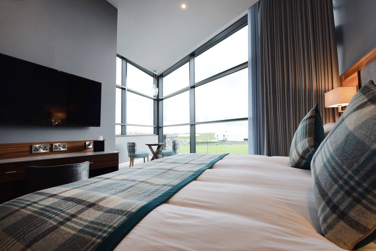 Quarto de hotel moderno com uma cama grande, uma TV de tela plana, uma escrivaninha com abajur e vista para um campo de golfe através de amplas janelas.