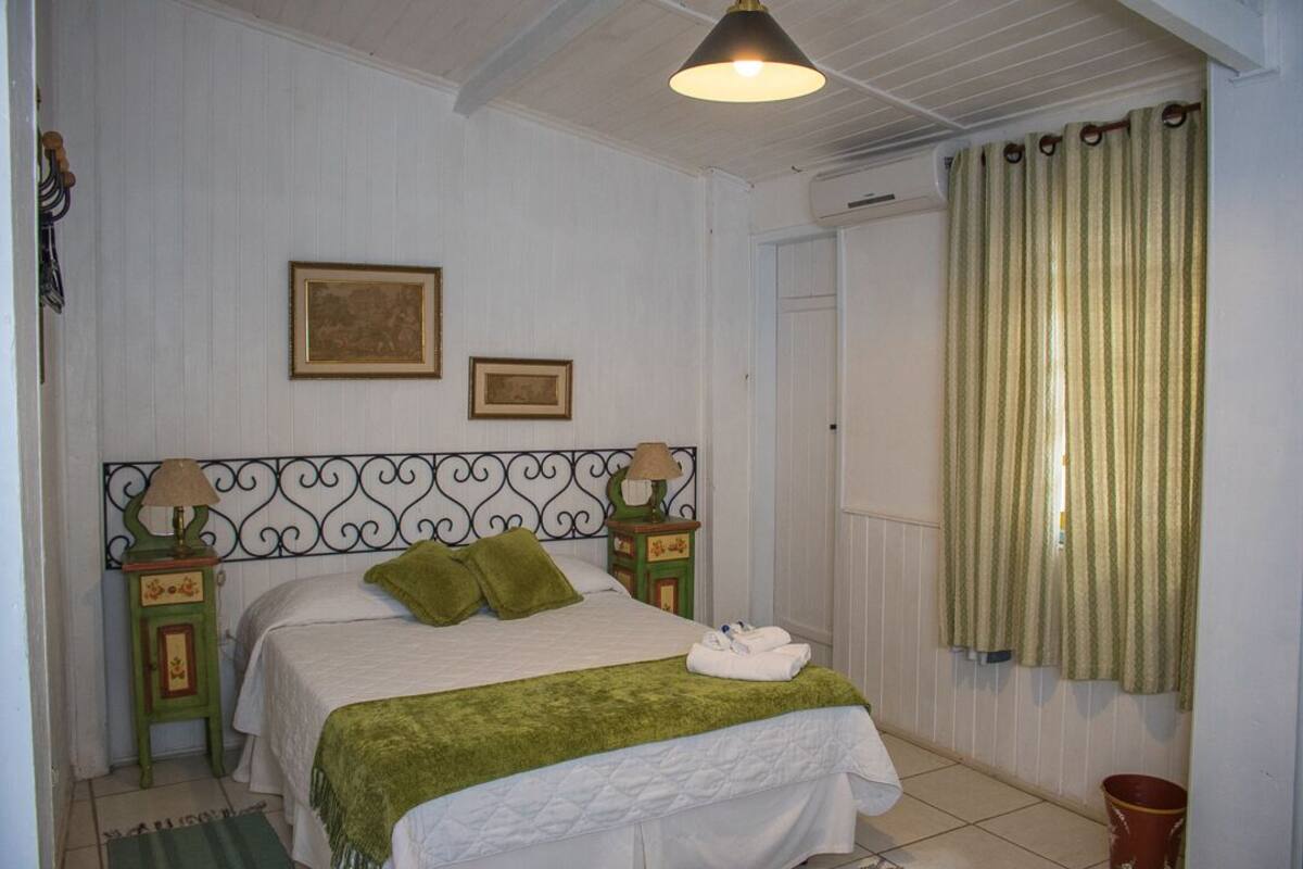 Une chambre à coucher avec un lit, deux tables de chevet, une lampe, un plaid vert et une fenêtre avec des rideaux.