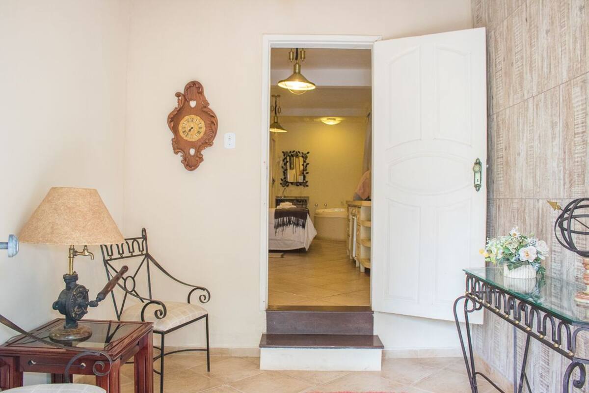 Un couloir avec une chaise en bois, une table avec une lampe, une horloge murale et une porte donnant sur une salle de bain équipée d’une baignoire et d’un miroir.