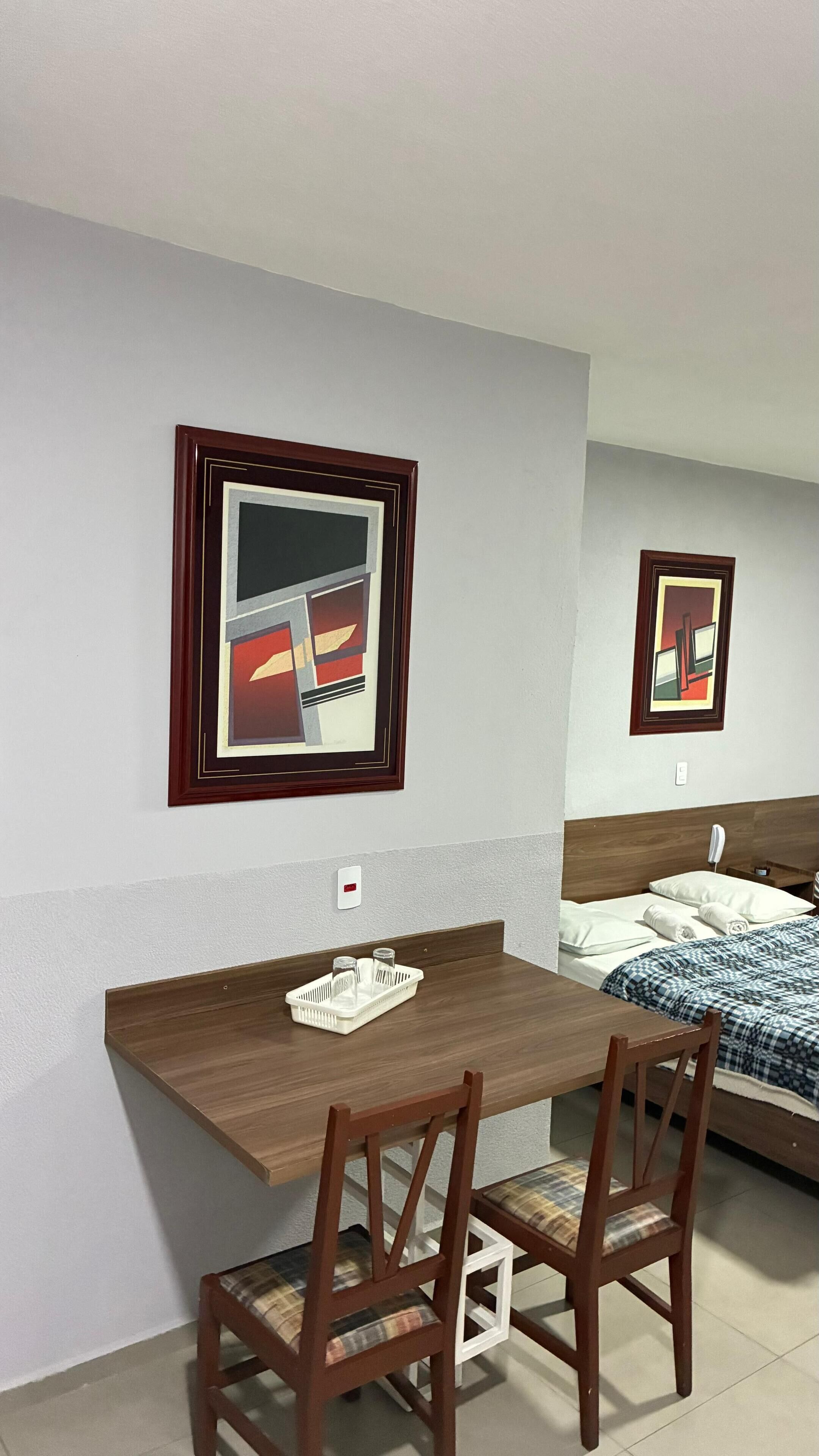 Habitación de hotel con un escritorio de madera y dos sillas, una cama con manta a cuadros azul y blanco, y dos cuadros abstractos en la pared.