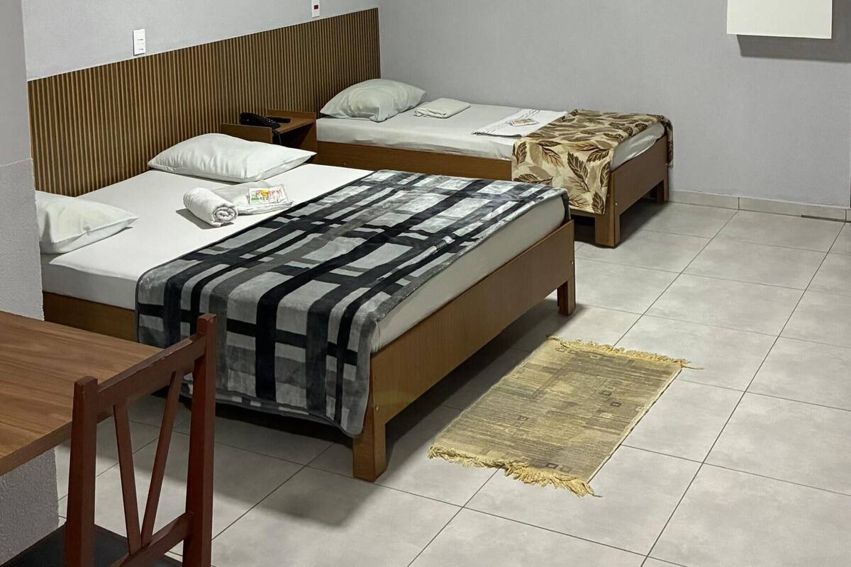 A small, clean room with two beds, a wooden chair, a table, and a wall-mounted air conditioner.