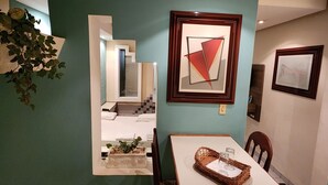 Un comedor con una mesa blanca, sillas de madera y una pared verde. Hay una obra de arte abstracta enmarcada sobre la mesa y una planta en maceta a la izquierda.
