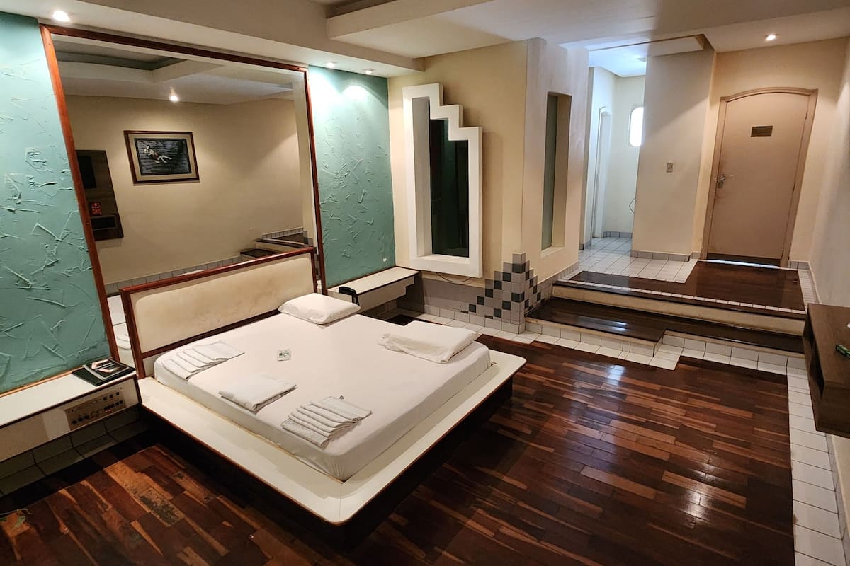 A bedroom with a large bed, wooden flooring, a mirror, and a door leading to another room.