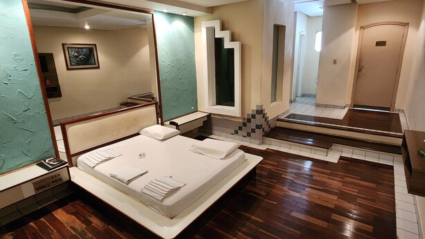 Un dormitorio con una cama grande, piso de madera, un espejo y una puerta que da a otra habitación.