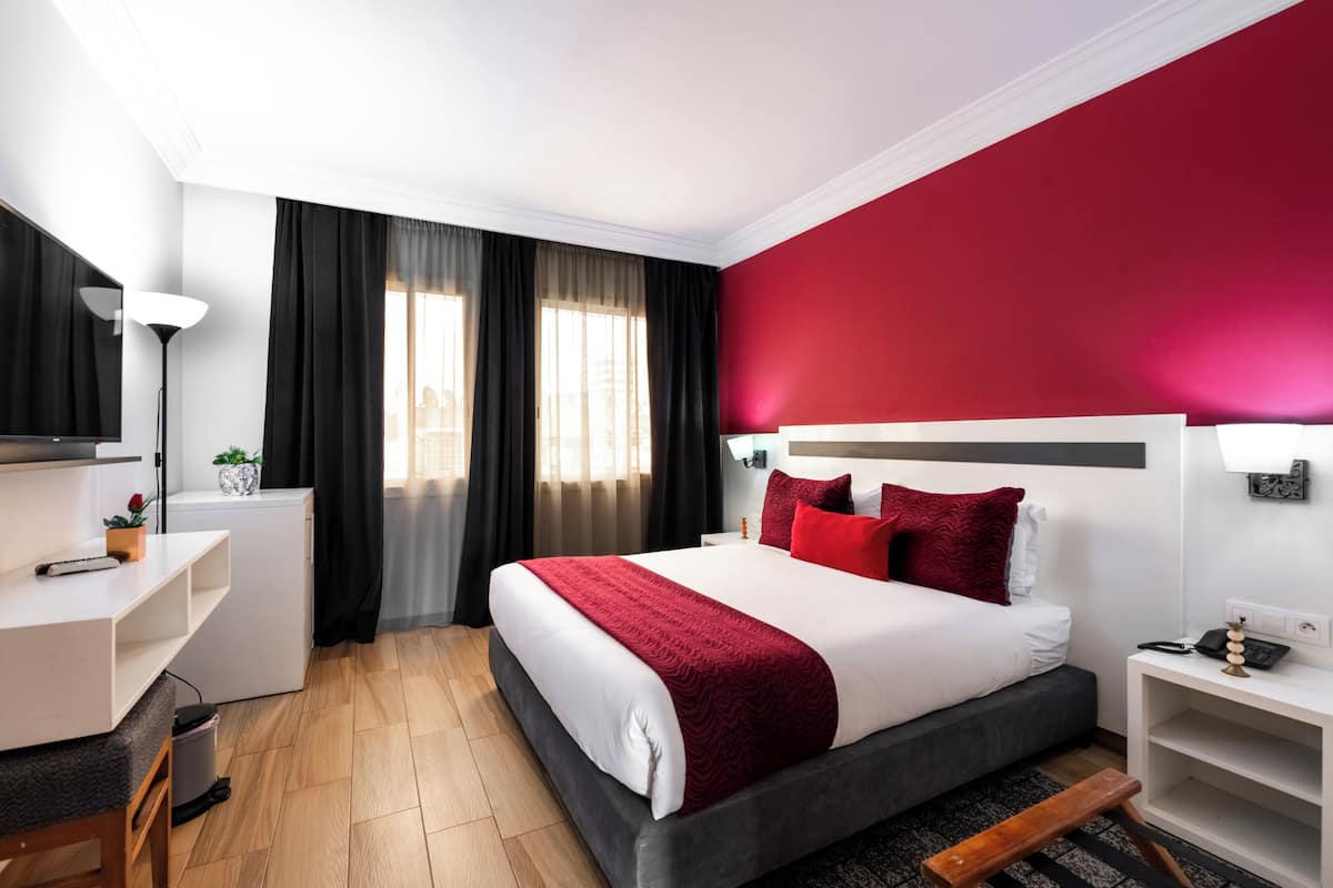A modern hotel room with a large bed, a desk, a chair, a small table, and a bedside lamp.