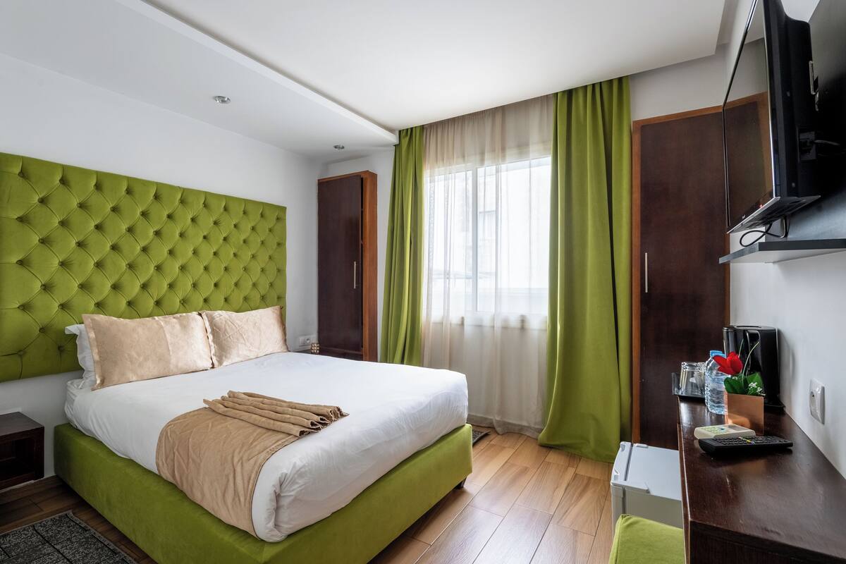 A hotel room with a green upholstered headboard, a large bed with white linens, a desk with a television, and a window with green curtains.