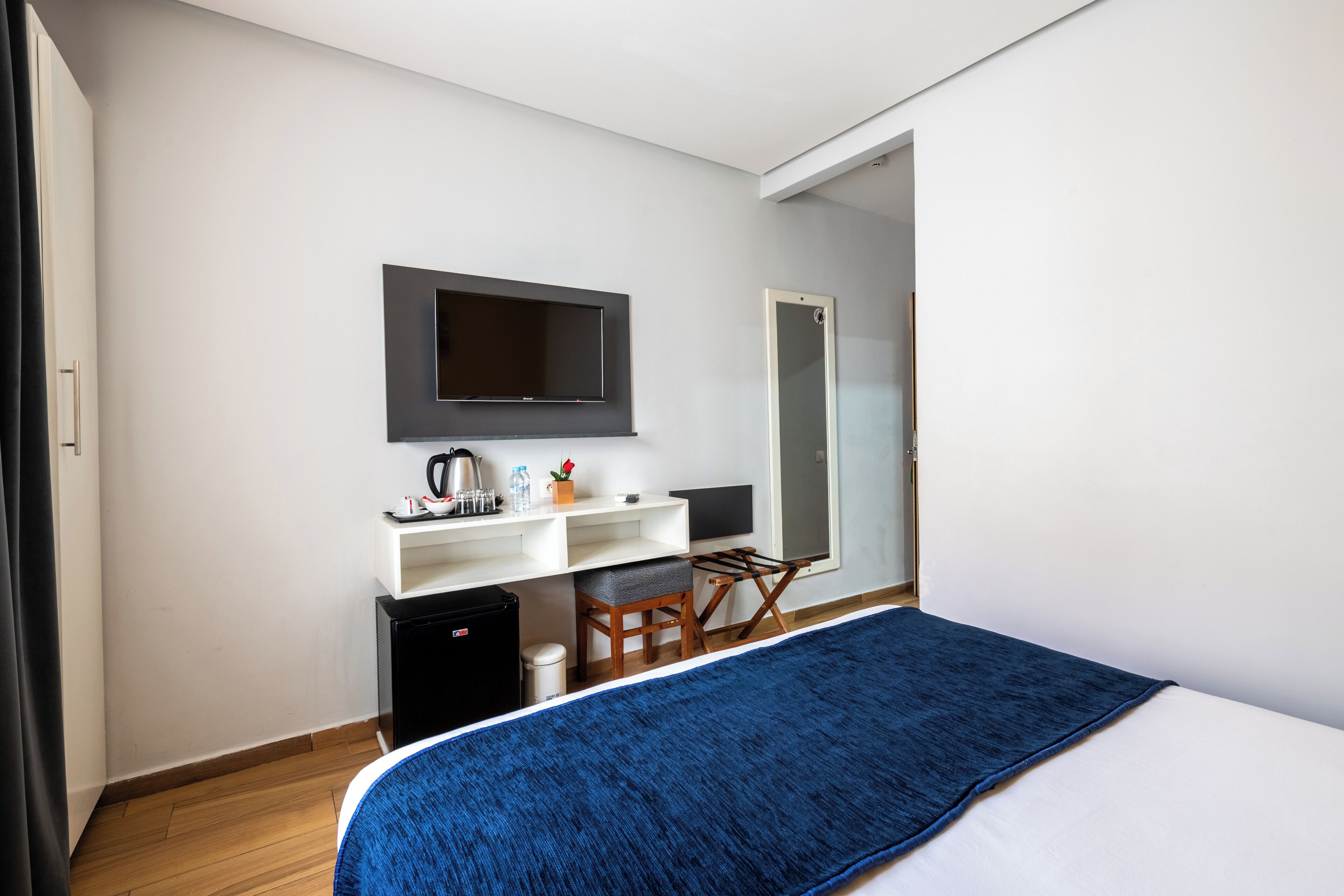 A modern hotel room with a bed, a desk with a flat-screen TV, a small refrigerator, and a mirror.