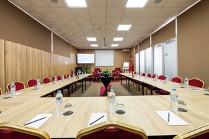 Un salón de conferencias con mesas largas, sillas rojas, botellas de agua y una pantalla de proyector.