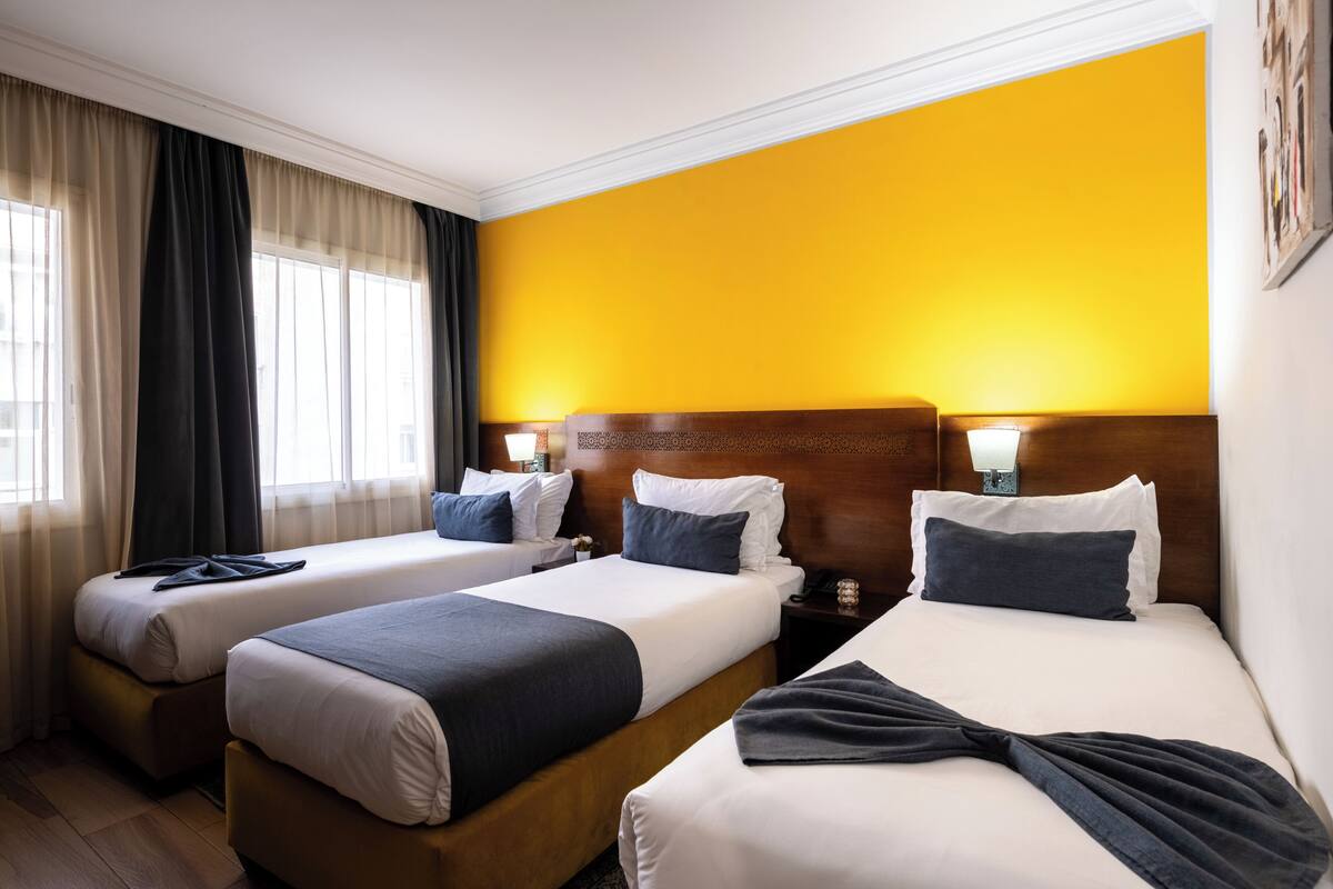 A hotel room with three beds, a wooden headboard, and a bright yellow accent wall.