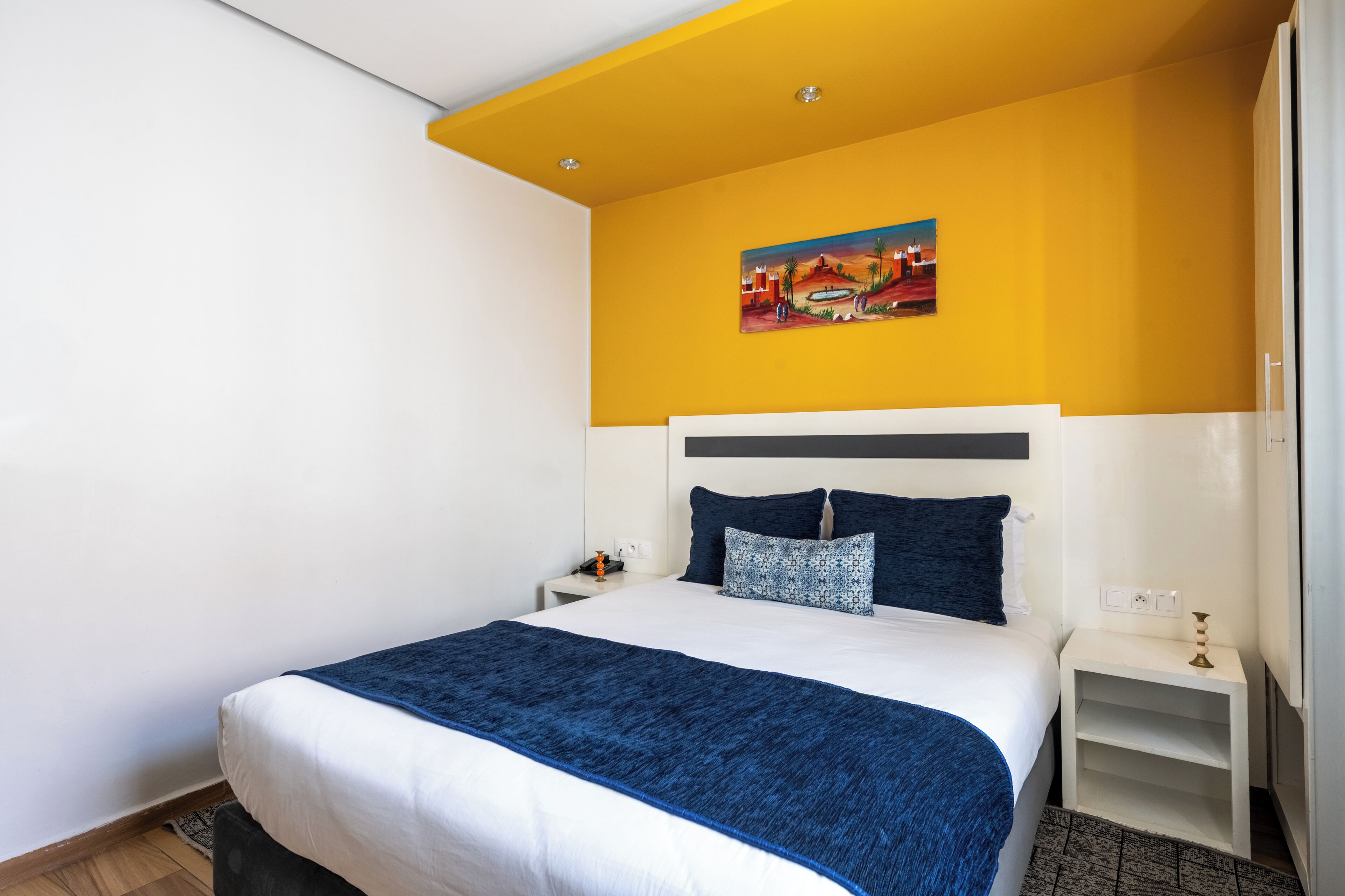 Une chambre d’hôtel avec un lit, du linge de lit blanc et bleu, une table de chevet, une petite table et un tableau au mur.