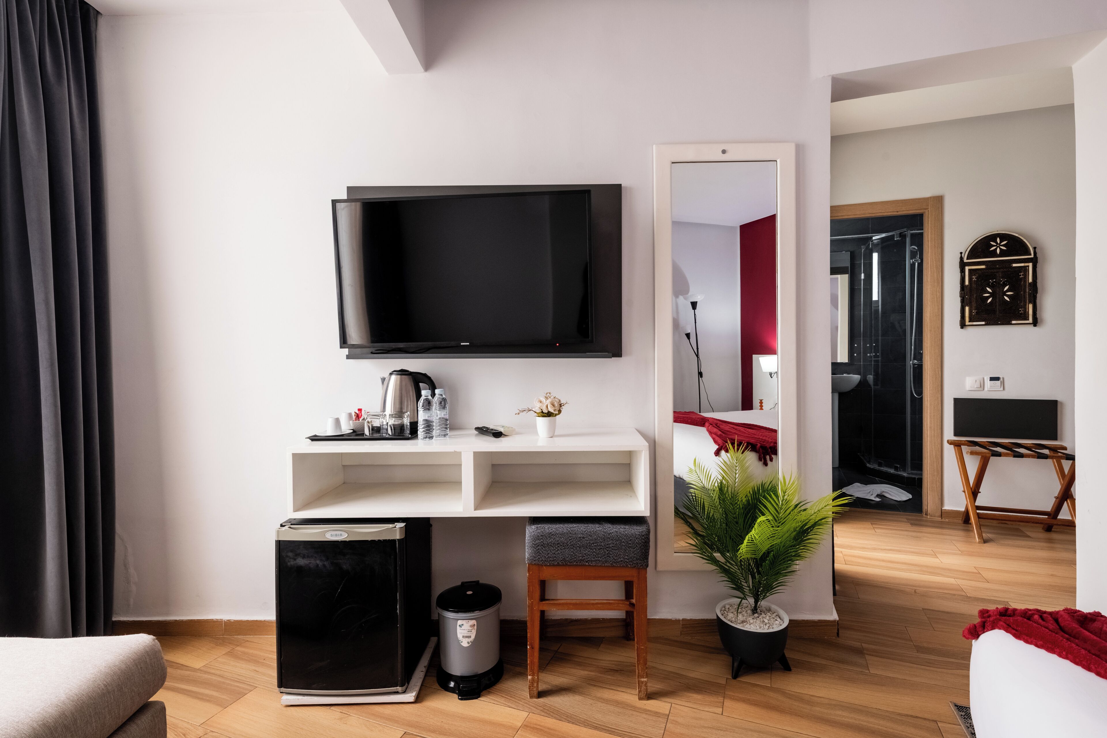 A modern living room with a flat-screen TV, a coffee machine, a small trash bin, a wooden stool, and a potted plant.